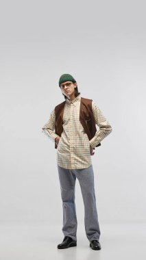 İşaretli gömlek giymiş, kahverengi yelek giymiş yeşil bereli ve gözlüklü sıradan kot pantolon giymiş stüdyonun arka planında poz veren genç bir adamın tam boy görüntüsü. Moda kavramı, gençlik kültürü, tarz, reklam