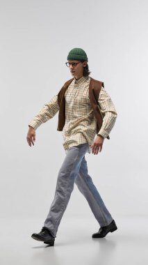 Yeşil bereli yakışıklı genç adam, gözlüklü, retro-stil tişörtlü ve kahverengi yelekli, hafif stüdyo arka planında poz veriyor. Moda kavramı, gençlik kültürü, şehir tarzı, reklam