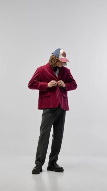 Kırmızı kadife ceket, gri pantolon ve kep giyen beyaz stüdyo arka planında poz veren kendinden emin bir gencin tam boy portresi. Cesur aksesuarlar. Moda kavramı, gençlik kültürü, şehir tarzı, reklam