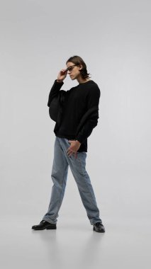 Rahatlamış özgüven. Siyah ceketli, kazaklı, açık kot pantolonlu ve gözlüklü beyaz stüdyo arka planında poz veren bir gencin tam boy portresi. Moda kavramı, gençlik kültürü, tarz, reklam