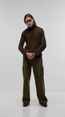 Moda gözlük takan, kahverengi örgü süveter ve pantolon giyen, ışık stüdyosunun arka planında poz veren yakışıklı bir gencin tam boy portresi. Moda kavramı, gençlik kültürü, şehir tarzı, reklam