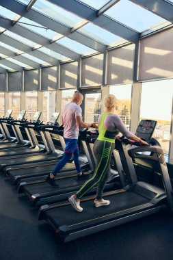 Spor giyim sektöründe yaşlı bir adam ve kadın koşu bandında yürüyor, modern spor salonunda antrenman yapıyor, sağlıklı ve aktif bir yaşam tarzı sürdürüyor. Yaşlıların konsepti, spor, aktif yaşam tarzı, sağlık