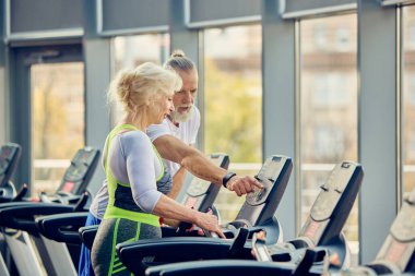 Yaşlı bir adam koşu bandındaki egzersizlerinde yaşlı bir kadına yardım ediyor, modern fitness salonunda ekipmanla antrenman yapıyor. Yaşlıların konsepti, spor, aktif yaşam tarzı, sağlık