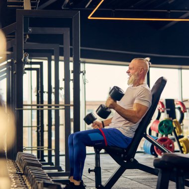 Atletik kıyafet eğitiminde son sınıf öğrencisi, iyi donanımlı spor salonunda dambılları kaldırıyor, yaşlılıkta dayanıklılık ve zindelik gösteriyor. Yaşlıların konsepti, spor, aktif yaşam tarzı, sağlık