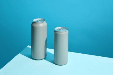 Soda, bira veya enerji içecekleri için iki gri alüminyum içecek kutusu, açık mavi zemin üzerinde canlı mavi arka plana karşı durarak, minimalist ve modern kompozisyon oluşturur. İçki markalaşması için model.