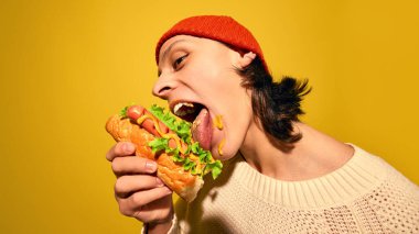 Kırmızı bereli genç adam portresi duygusal olarak marullu sosisli yiyor, hardal yalıyor, parlak sarı arka planda duruyor. Sokak yemeği kavramı, kış, pop sanat, fuarlar, duygular