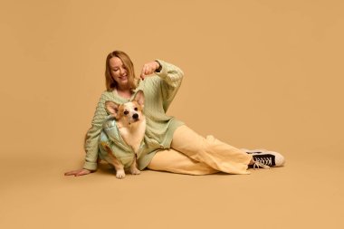 Güzel, açık yeşil hırka giyen, poz veren, stüdyo arka planında eşleşen yeşil ceket giyen sevimli Corgi köpeğiyle oynayan genç bir kadın. Hayvan, kış mevsimi, arkadaşlık konsepti.