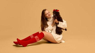 Örgü süveter ve Red Sox giyen genç bir kadın. Stüdyoda çalışan sevimli siyah kaniş köpeğiyle poz veriyor. Hayvan kavramı, kış mevsimi, arkadaşlık, mutluluk.