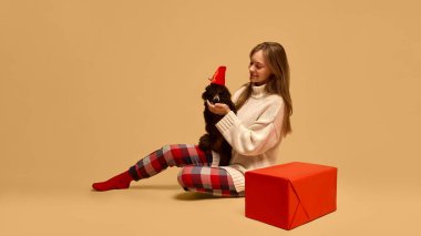 Gelmiş geçmiş en iyi hediye. Sıcak kış kıyafetleri ve damalı pijama pantolonu giymiş, kırmızı Noel şapkalı sevimli fino köpeğiyle poz veren genç bir kadın. Hayvan kavramı, kış mevsimi, arkadaşlık, mutluluk.