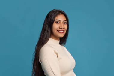 Siyah saçlı, uzun kollu, mavi stüdyo arka planına gülümseyen güzel bir Hintli kadının yan görüntüsü. İnsan duyguları kavramı, yüz ifadesi, gençlik.