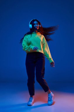 Genç Hintli bir kadının günlük giysiler içindeki portresi neon ışıkta mavi arka planda kulaklıkla müzik dinliyor. İnsan duyguları kavramı, yüz ifadesi, gençlik.