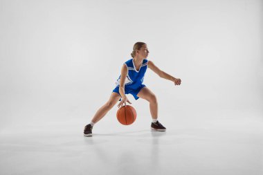 Rekabetçi genç kız, mavi üniformalı basketbolcu topla hareket halinde, beyaz stüdyo geçmişine karşı kazanmak için motivasyonlu antrenman yapıyor. Spor anlayışı, gençlik, hareket, sağlıklı yaşam tarzı