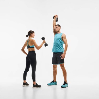 Dambıl ve kaslı adamla birlikte beyaz stüdyo arka planına karşı kettlebell ile el egzersizi yapan zayıf genç kadın eğitimi. Spor anlayışı, aktif ve sağlıklı yaşam tarzı, egzersiz, fitness
