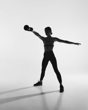 Siyah beyaz fotoğrafçılık. İnce vücutlu, eğitimli, kettlebell ile egzersiz yapan genç bir kadının tam boy görüntüsü. Spor anlayışı, aktif ve sağlıklı yaşam tarzı, egzersiz, fitness