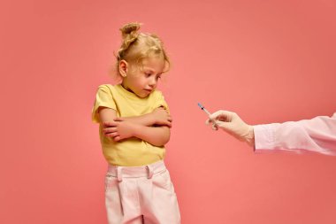 Küçük kız, çocuk korku içinde kollarını kavuşturuyor, gergin ve korkmuş görünüyor doktor pembe stüdyo arka planına aşı yaparken. Sağlık, tıbbi tedavi, çocukluk kavramı