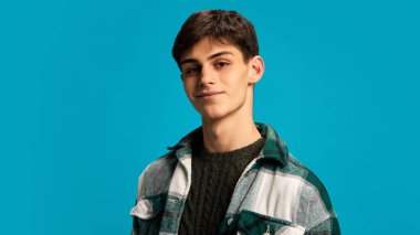 Siyah tişört ve kareli ceket giyen, mavi stüdyo arka planında küçük bir gülümsemeyle poz veren, koyu renk saçlı genç bir adamın portresi. Gençlik kavramı, duygular, yaşam tarzı, moda