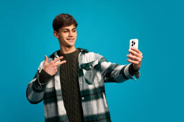 Günlük giysiler içinde yakışıklı bir genç, kareli tişörtlü akıllı telefonlu özçekimini mavi stüdyo geçmişine karşı yapıyor. Sosyal medya, iletişim. Gençlik kavramı, duygular, yaşam tarzı, moda