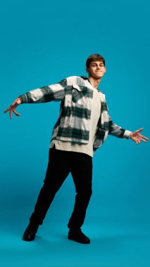 Yakışıklı bir genç anne, günlük kıyafetler giymiş, mavi stüdyo arka planında neşeyle dans eden kareli gömlek. Gençlik kavramı, duygular, yaşam tarzı, moda, pozitif hava