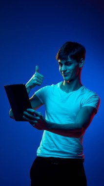 Yakışıklı, gülümseyen bir adam, videoyla tabletle konuşan bir öğrenci, baş parmağını kaldırıyor, mavi ışıkta mavi stüdyonun arka planına karşı olumlu bir hareket yapıyor. Gençlik kavramı, duygular, yaşam tarzı, iletişim