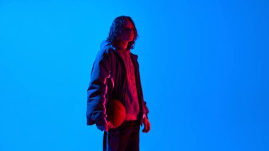 Uzun kıvırcık saçlı, geniş spor giysiler giymiş, mavi arka planda basketbolla ayakta duran yakışıklı bir genç portresi. Spor, gençlik, tarz, modern moda konsepti