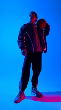 Şık spor kıyafetleri içinde, basketbolla birlikte neon ışıklarıyla arka planda poz veren yakışıklı bir gencin tam boy düşük açılı portresi. Spor anlayışı, gençlik, sokak tarzı, moda