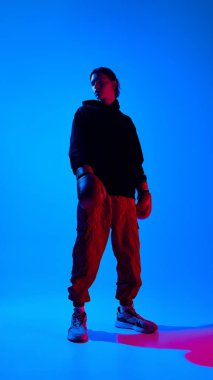 Spor giyimli, boks eldivenli, mavi arka planda neon ışıkta duran yakışıklı bir gencin geniş açılı görüntüsü. Spor anlayışı, gençlik, sokak tarzı, modern moda.