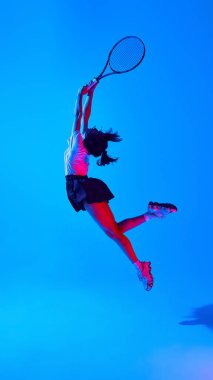 Tişört ve etek giymiş, tenis raketiyle hareket eden, mavi arka planda neon ışığı altında genç ve şık bir kadının tam boy dinamik görüntüsü. Spor anlayışı, gençlik, sokak tarzı, modern moda.