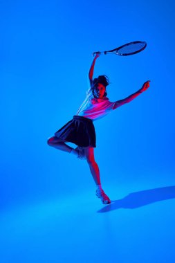 Gömlekli ve etekli genç bir kadının dinamik görüntüsü. Tenis raketiyle neon ışıkta mavi stüdyo arka planına karşı poz veriyor. Spor anlayışı, gençlik, sokak tarzı, modern moda.