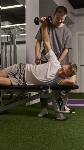 Мужчина лежит на боку на тренажерке, выполняя упражнения по реабилитации плеч с гантелями, при содействии тренера. Концепция реабилитации, восстановления, здравоохранения, физической терапии