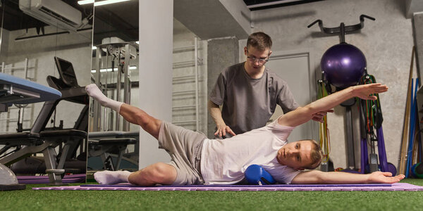 Мужчина растягивается на коврике для йоги с тренерской помощью, используя ролики для реабилитации позвоночника в яркой спортивной среде. Концепция реабилитации, восстановления, здравоохранения, физической терапии