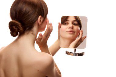 Arka planda, ayna tutan ve beyaz stüdyo arka planına nemlendirici krem süren güzel bir kadın resmi var. Doğal güzellik, cilt bakımı, kozmetik ve kozmetik kavramı