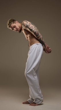 Üstsüz, atletik, dövmeli, gri spor pantolonlu, tarafsız stüdyo geçmişli yakışıklı bir gencin tam boy portresi. Erkek güzelliği, moda, vücut bakımı, spor kavramı