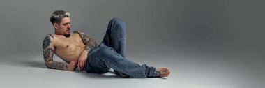 Gömleksiz, kaslı dövmeli, yerde oturan, kot pantolonla poz veren yakışıklı genç adam gri stüdyo arka planına sahip. Erkek güzelliği, moda, vücut bakımı, spor anlayışı. Pankart. İlan için alanı kopyala