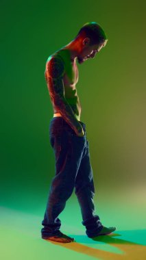 Uzun boylu, şık saç stili olan, kotsuz poz veren, neon ışıklı yeşil arka planlı yakışıklı bir genç. Gömleksiz dövmeli vücut. Erkek güzelliği, moda, vücut bakımı kavramı