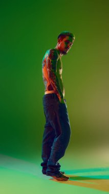 Uzun boylu, şık saç stili olan, kotsuz poz veren, neon ışıklı yeşil arka planlı yakışıklı bir genç. Gömleksiz dövmeli vücut. Erkek güzelliği, moda, vücut bakımı kavramı