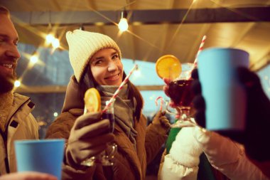Kışın sıcak giysiler içinde gülümseyen genç adam ve kadın kış akşamını dışarıda sıcak şarap içerek, sıcak içecekler içerek geçiriyorlar. Kış tatili kavramı, Noel, gelenekler, etkinlik