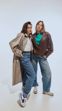 Trençkotlu şık genç kadınlar, büyük beden ceket ve geniş bacaklı kotlar, beyaz stüdyo arka planında eski moda kıyafetlerle poz veriyorlar. 90 'ların modası, eski moda, gençlik kavramı