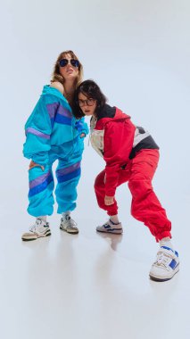 90 'ların ikonik rüzgarlığı ve koyu renkli eşofmanlarıyla beyaz stüdyo arka planında şık bir poz veren iki genç kadın. 90 'ların modası, eski moda, gençlik kavramı