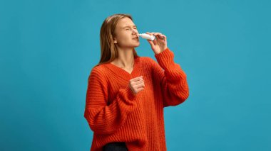 Mavi stüdyo arka planında burun spreyi kullanan turuncu kazaklı genç bir kadın. Mevsimsel alerji, grip ve sinüs enfeksiyonu. Hastalık, ilaç, tedavi, sağlık