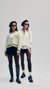 Örgü yelek ve hırka giyen, başlık takan, güneş gözlüğü takan ve stüdyo arka planında modaya uygun ayakkabılar giyen iki genç kadının tam boy portresi. İş anlayışı, moda eğilimleri, tarz