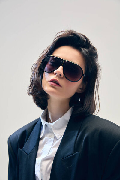 Portrait of stylish and beautiful young woman with oversized sunglasses, black blazer and white shirt, posing against minimalist studio background. Concept of business, fashion trends, beauty, style