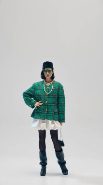 Sonsuz zarafet. Yeşil ceketli, beyaz gömlekli, kadife bereli, güneş gözlüklü ve stüdyo arka planında poz veren çekici genç bir kadın. İş anlayışı, moda eğilimleri, güzellik, tarz