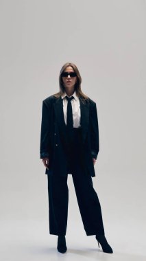 Zarif ve şık bir iş kadını, beyaz gömlek, kravat ve güneş gözlükleriyle tamamen siyah bir takım giyer ve stüdyo geçmişine karşı özgüvenle poz verir. İş anlayışı, moda eğilimleri, güzellik, tarz