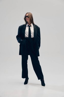 Üniformayla kendine güveni sağlamak. Siyah takım elbiseli, büyük beden ceketli, beyaz gömlekli, kravatlı ve güneş gözlüklü stüdyo arka planında poz veren bir kadın. İş anlayışı, moda eğilimleri