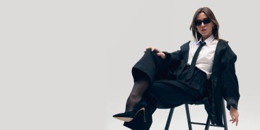 Kendinden emin ve rahat bir hava. Siyah takım elbiseli, beyaz gömlekli, kravatlı, güneş gözlüklü ve botlu şık bir kadın sandalyede oturuyor ve stüdyo arka planında poz veriyor. İş anlayışı, moda eğilimleri, güzellik, tarz