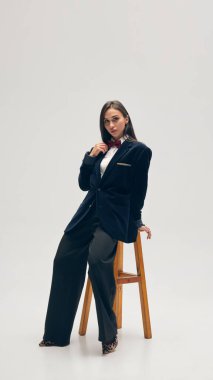 Kendine güvenen genç bir kadının tam boy portresi beyaz stüdyo arka planında kadife ceket, papyon, pantolon ve beyaz gömlek giyer. İş anlayışı, moda eğilimleri, güzellik, tarz