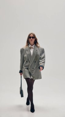 Uzun boylu, kendine güvenen, büyük gri ceketli, siyah taytlı, topuklu ve güneş gözlüklü, stüdyo arka planında küçük bir çantayla yürüyen bir kadın. İş anlayışı, moda eğilimleri, güzellik, tarz