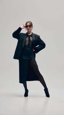 Kendine güveni tam bir iş kadını, aşırı büyük spor ceket, bluz, etek ve güneş gözlüğü açık gri stüdyo arka planında poz veriyor. İş anlayışı, moda eğilimleri, güzellik, tarz