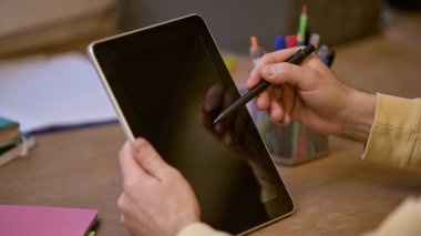 Tablet ve stilini tutan ellerin yakın çekimi, organize olmuş masa düzeniyle boş ekranı işaret ediyor, renkli kırtasiye malzemesi ve not defterleri var. İş kavramı, modern teknolojiler, dijital sanat