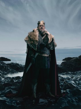 Sanat kolajı. Ortaçağ kralı, kürklü pelerinli ve zırhlı dramatik bulutlu gökyüzünün altında kayalık sahil şeridinde duruyor. Tarih kavramı, kraliyet, güç, fantezi